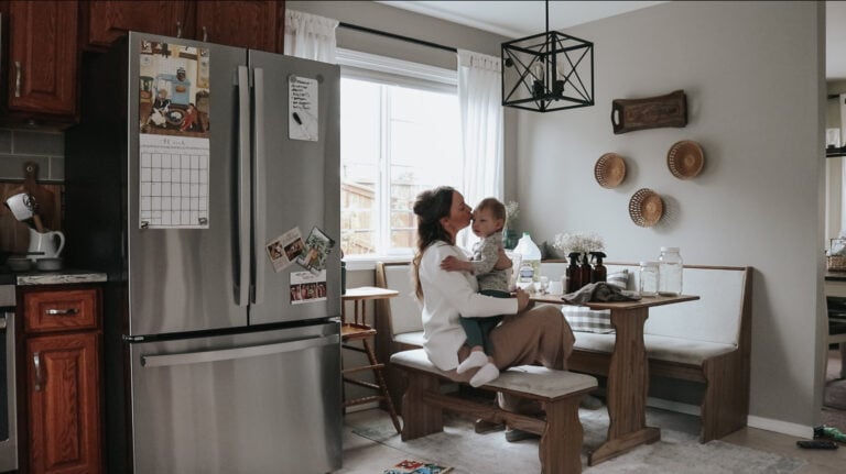 Homemaker sits at her kitchen table holding her child. She is kissing her child on the cheek.
